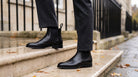 Brecon Chelsea Boot in black calf, worn with charcoal trousers on a stone step